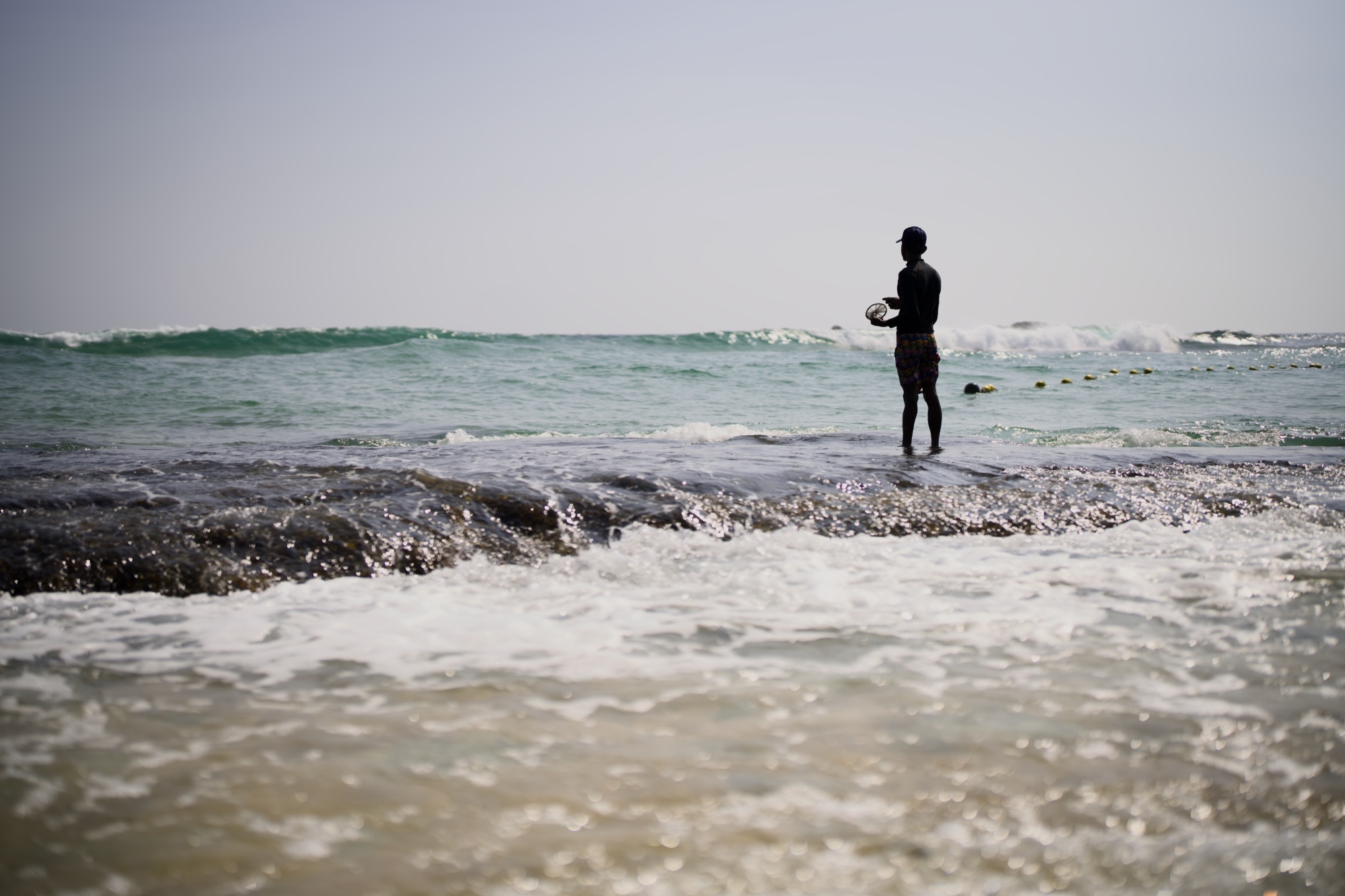 A fisherman standing on the rock with heave waves splashing him. Only his shadow is visible. He uses very rudimentary equipment.